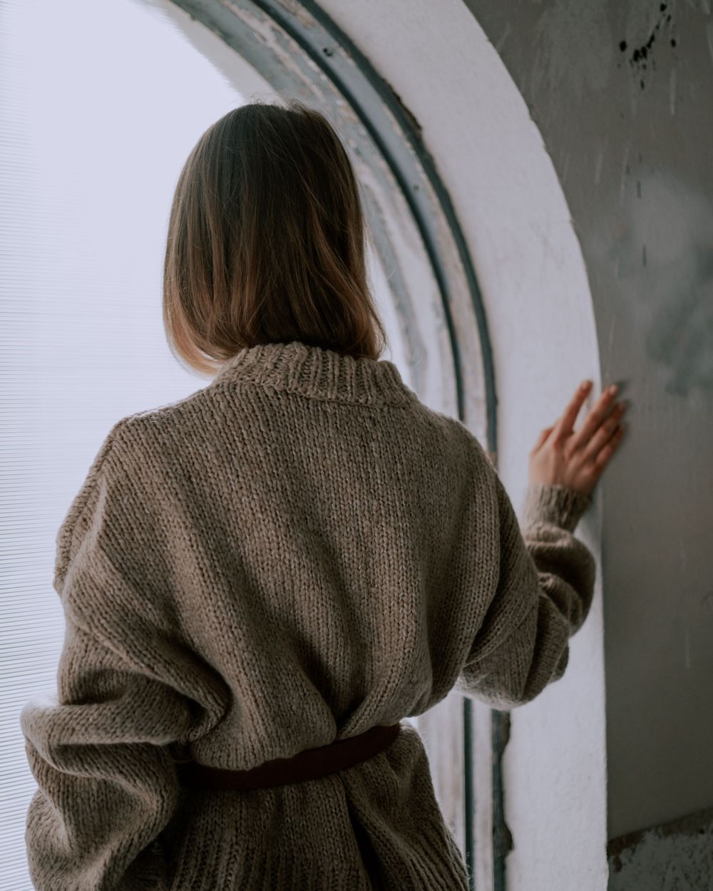 Mary Ellis, seen from behind in a knitted sweater, one hand on an arched window.