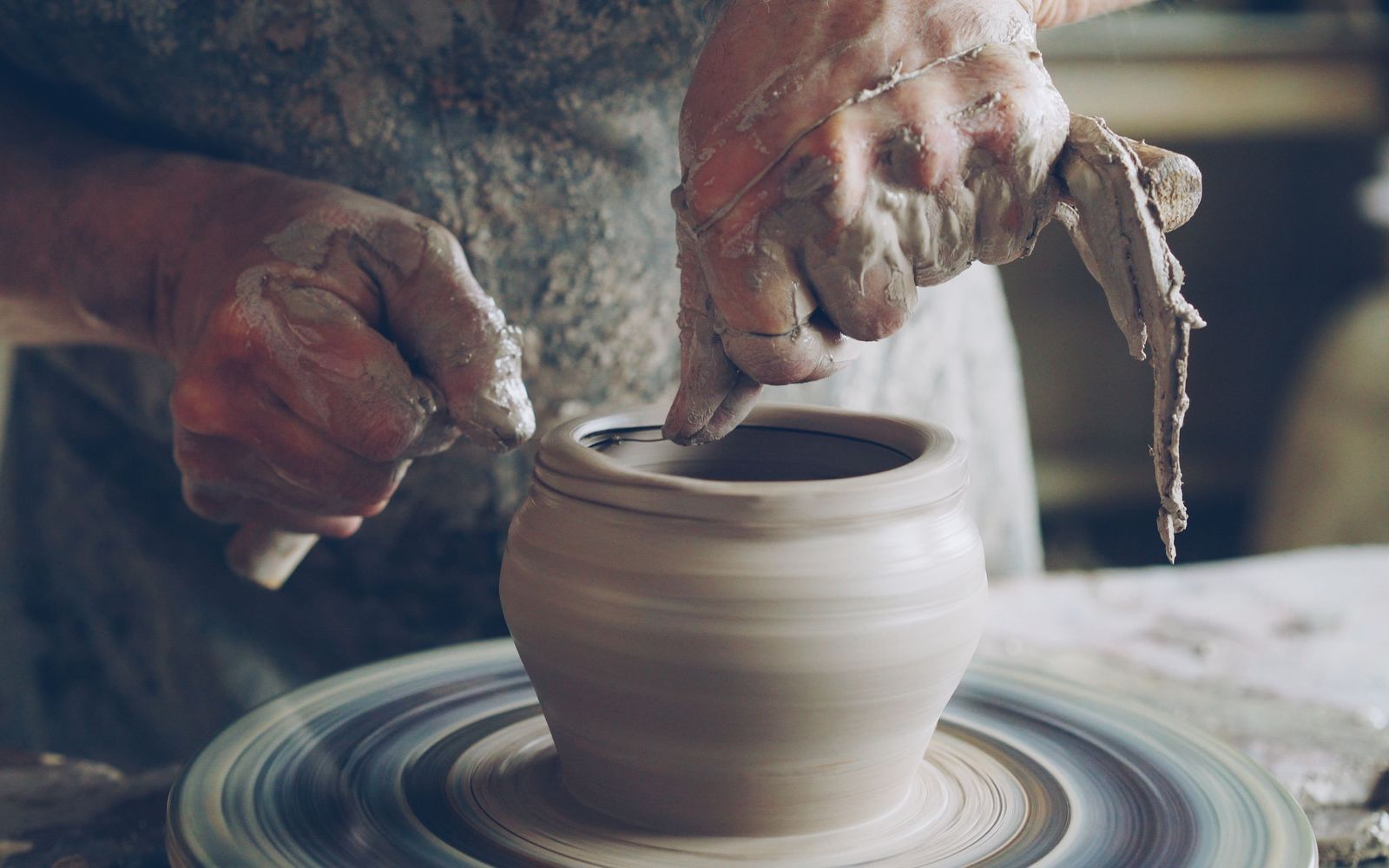 Clay-covered hands shaping a small pot on a spinning potter’s wheel.