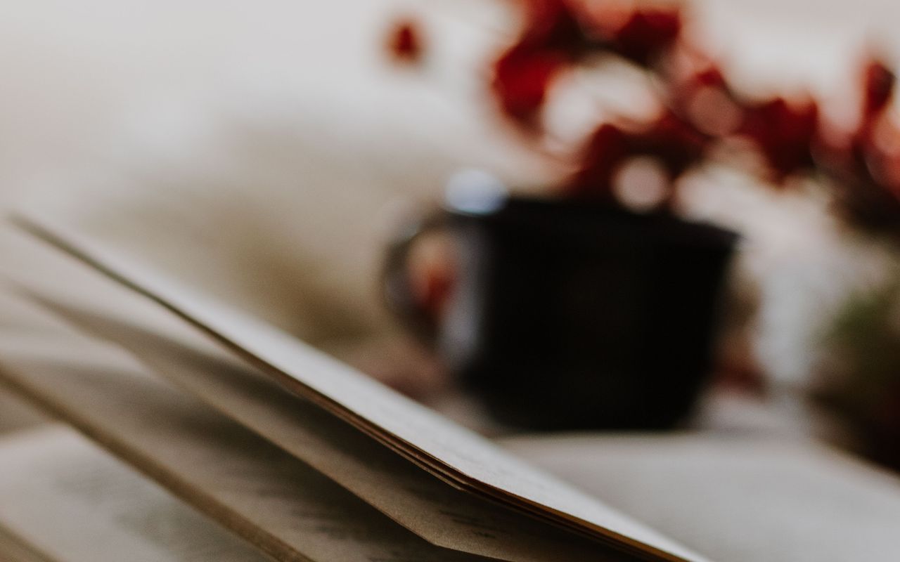An open book on a table, with a dark cup and red flowers soft in the background.